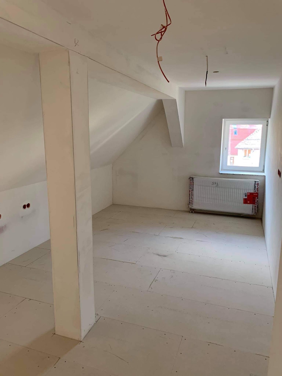 Poddasze w trakcie prac wykończeniowych, widoczne płyty na podłodze, otynkowane ściany, grzejnik z folią ochronną i wystające kable.