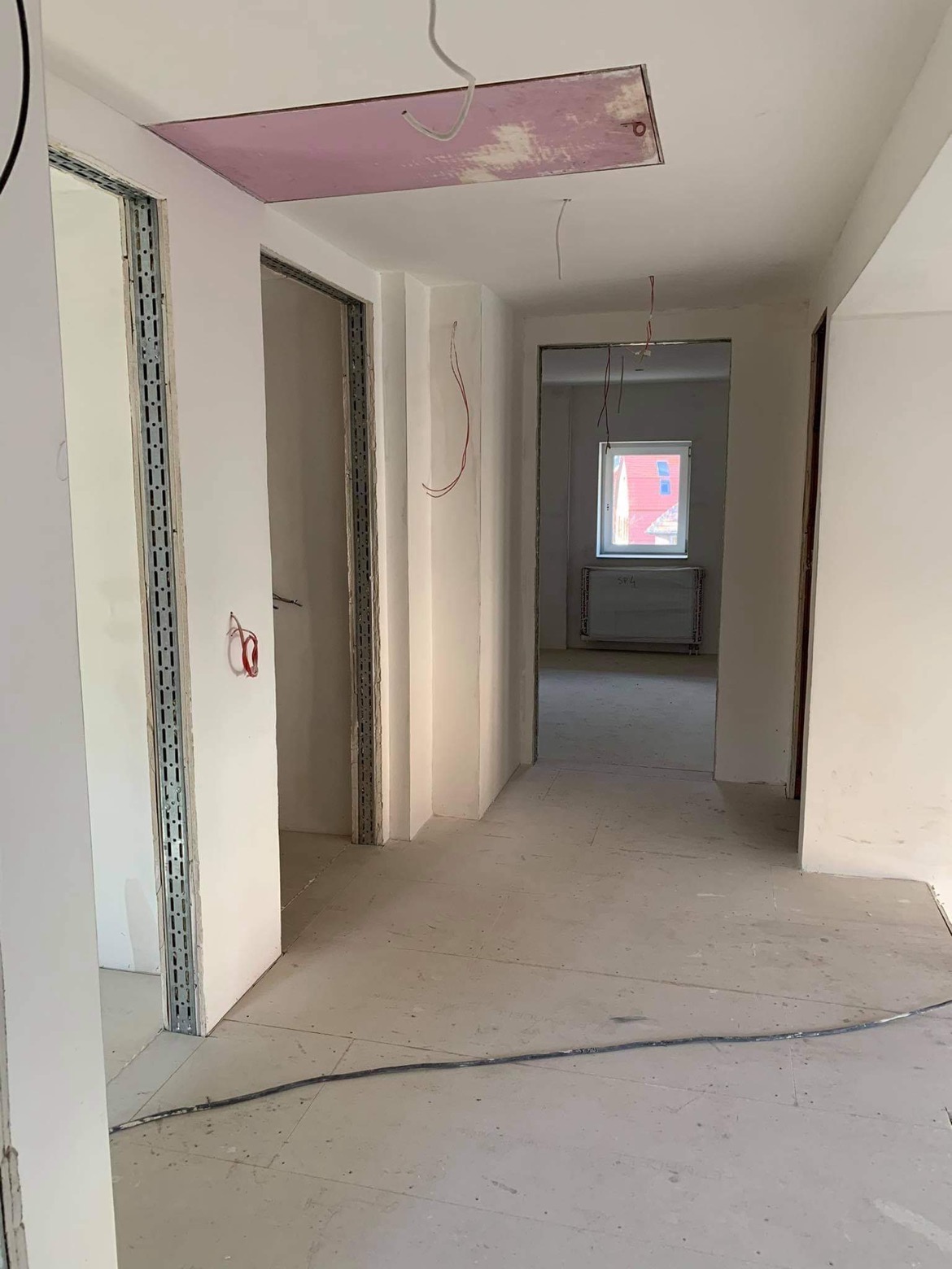 Wnętrze w trakcie remontu: surowe ściany po gładziowaniu, metalowe profile ościeżnic, widoczne kable elektryczne i fragment różowej płyty na suficie.