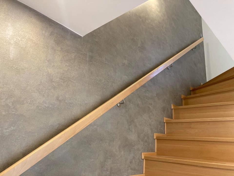 Schody drewniane z jasną poręczą na tle ściany pokrytej szarą dekoracyjną fakturą imitującą kamień.