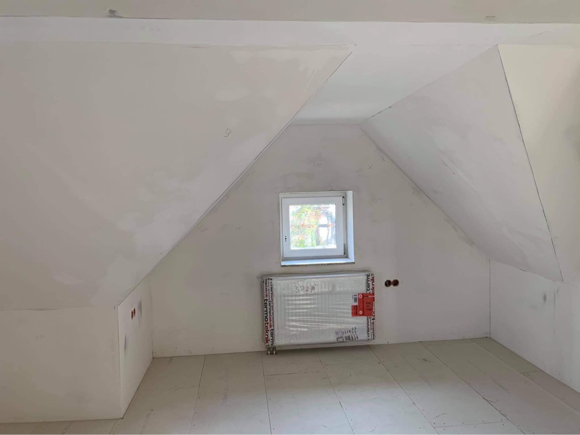 Poddasze w trakcie wykańczania: gładzie na skosach, okno w białej ramie, grzejnik w folii ochronnej, przygotowana podłoga.