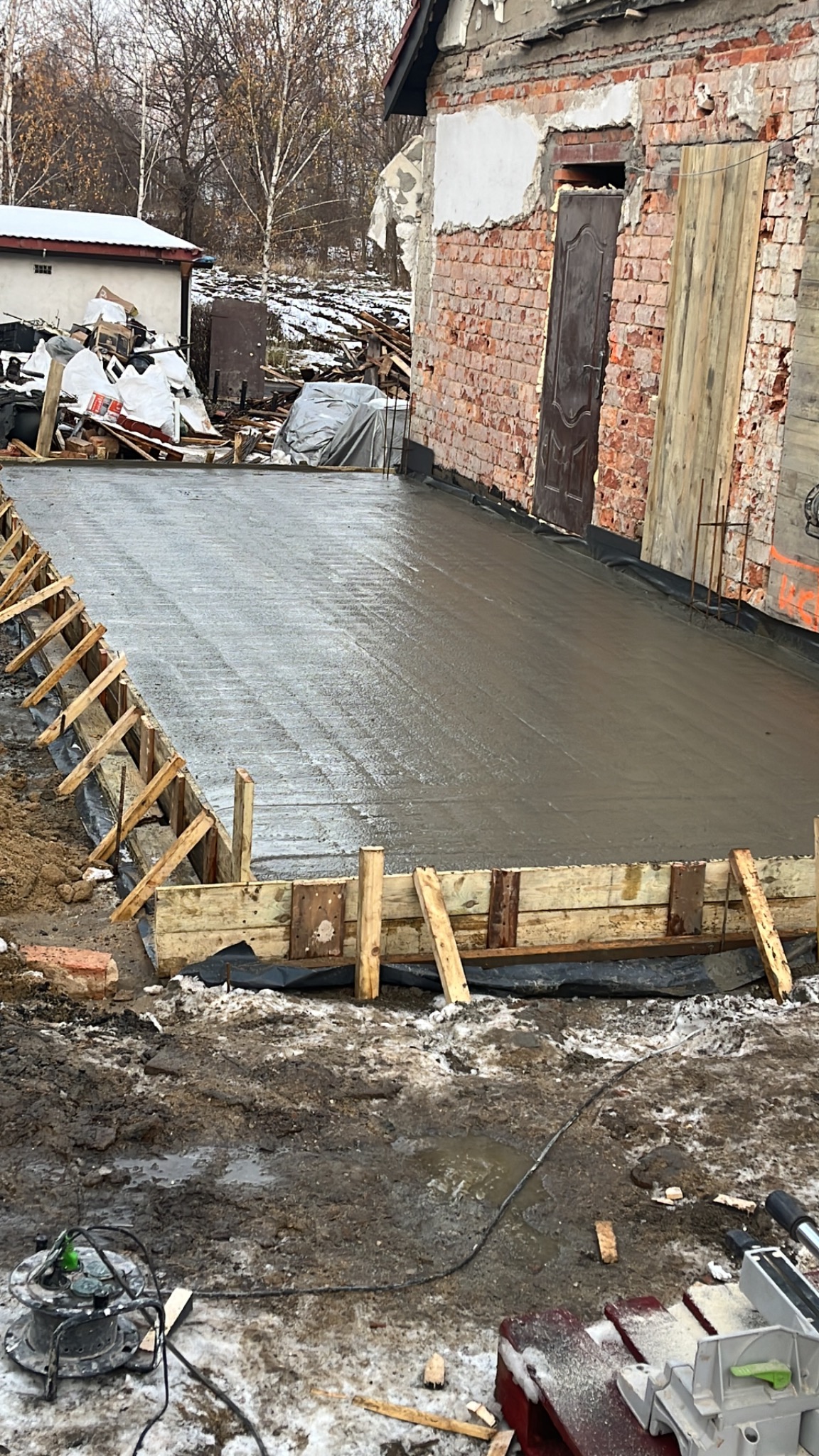 Świeżo wylany beton na podbudowie z drewnianym szalunkiem, widoczny fragment budynku z cegły i gruzu w tle, teren budowy z błotem i resztkami śniegu.