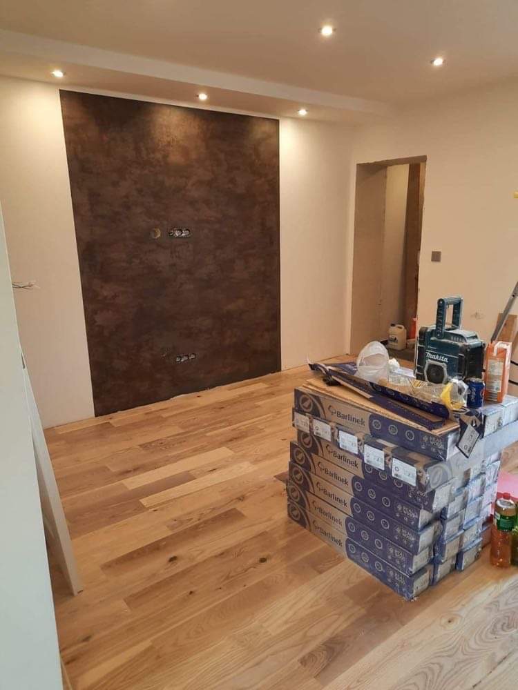 Pokój w trakcie wykańczania: świeżo położona drewniana podłoga Barlinek, ściana z dekoracyjną, brązową fakturą, widoczne puszki elektryczne, narzędzia i materiały budowlane.