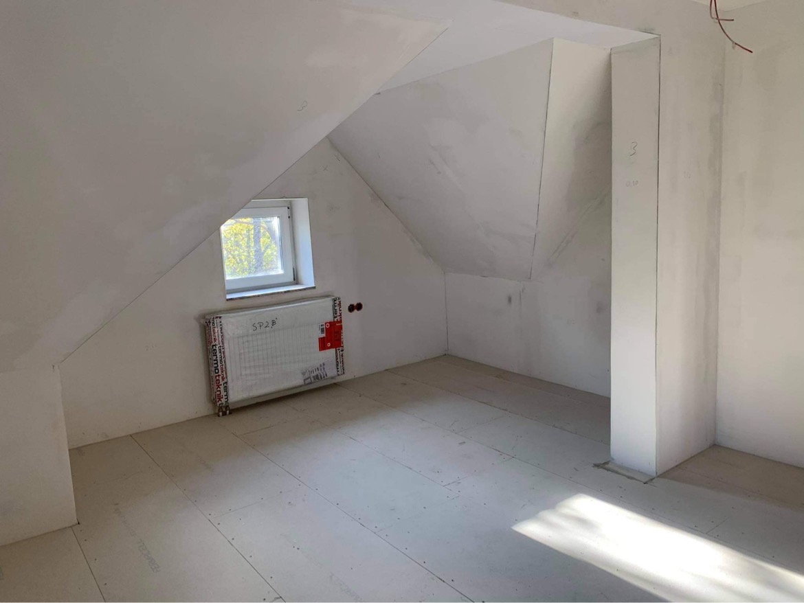 Poddasze ze skośnym dachem w trakcie prac wykończeniowych, ściany pokryte gładzią, widoczny grzejnik z folią ochronną, podłoga wyłożona płytami OSB, małe okno w szczycie dachu.