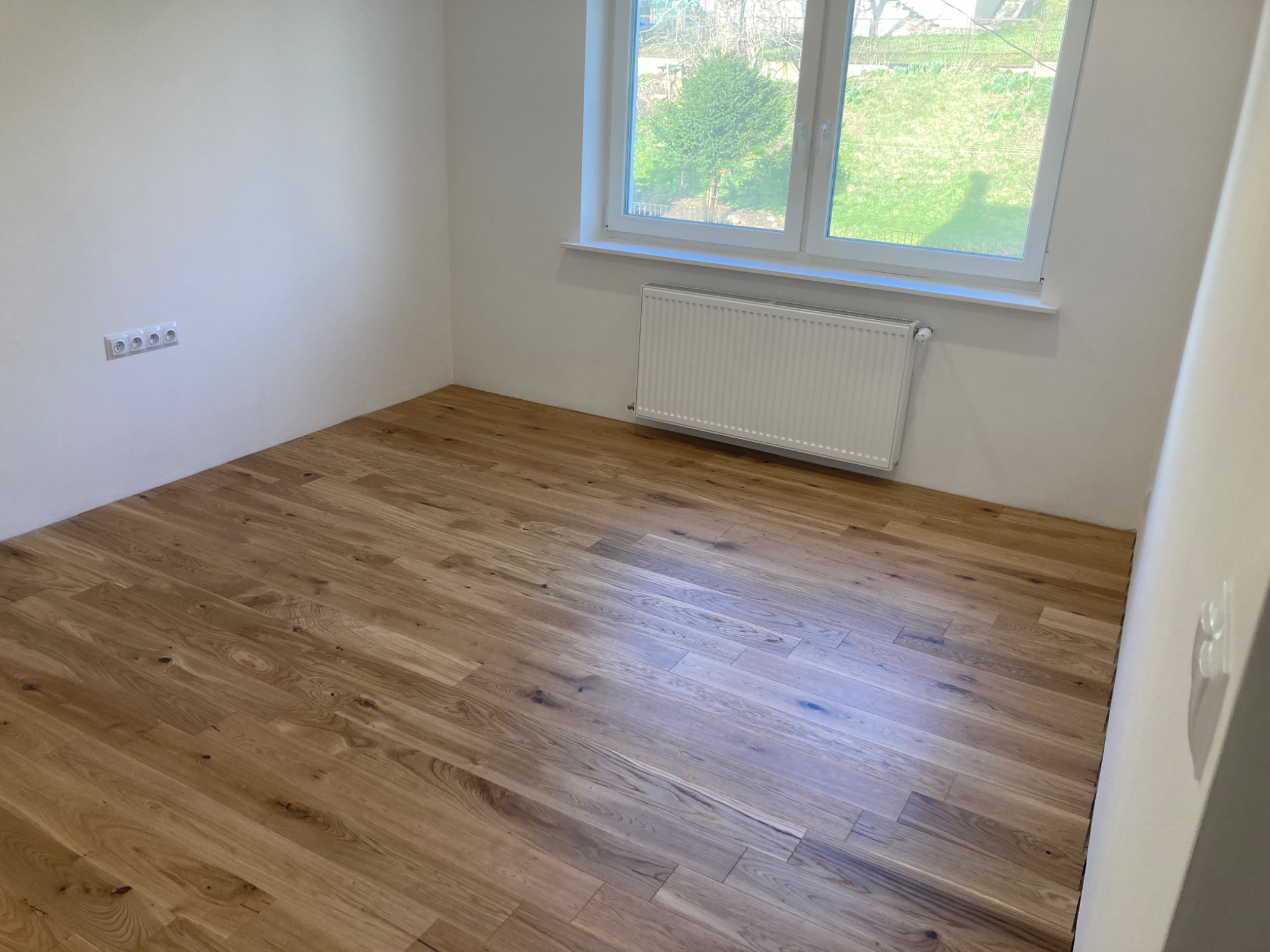 Pusty pokój po remoncie: świeżo położona drewniana podłoga, białe ściany z gniazdkami elektrycznymi, grzejnik pod oknem z widokiem na zieleń.