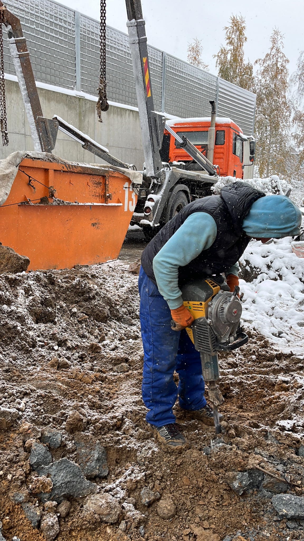 Pracownik w niebieskim ubraniu i pomarańczowych rękawicach używa młota pneumatycznego do rozbijania ziemi, obok stoi pomarańczowa ciężarówka z dźwigiem podczas lekkiego opadu śniegu.