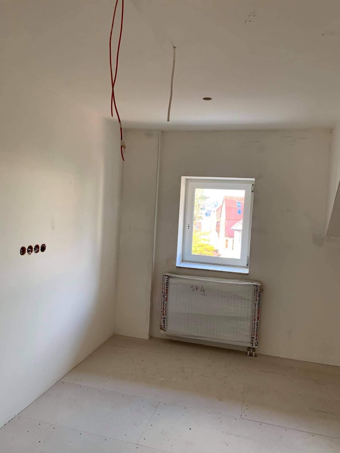 Surowe wnętrze pokoju w trakcie prac wykończeniowych: ściany po gładziowaniu, widoczne kable elektryczne, okno z widokiem na budynek, grzejnik zabezpieczony folią, podłoga przygotowana pod dalsze...