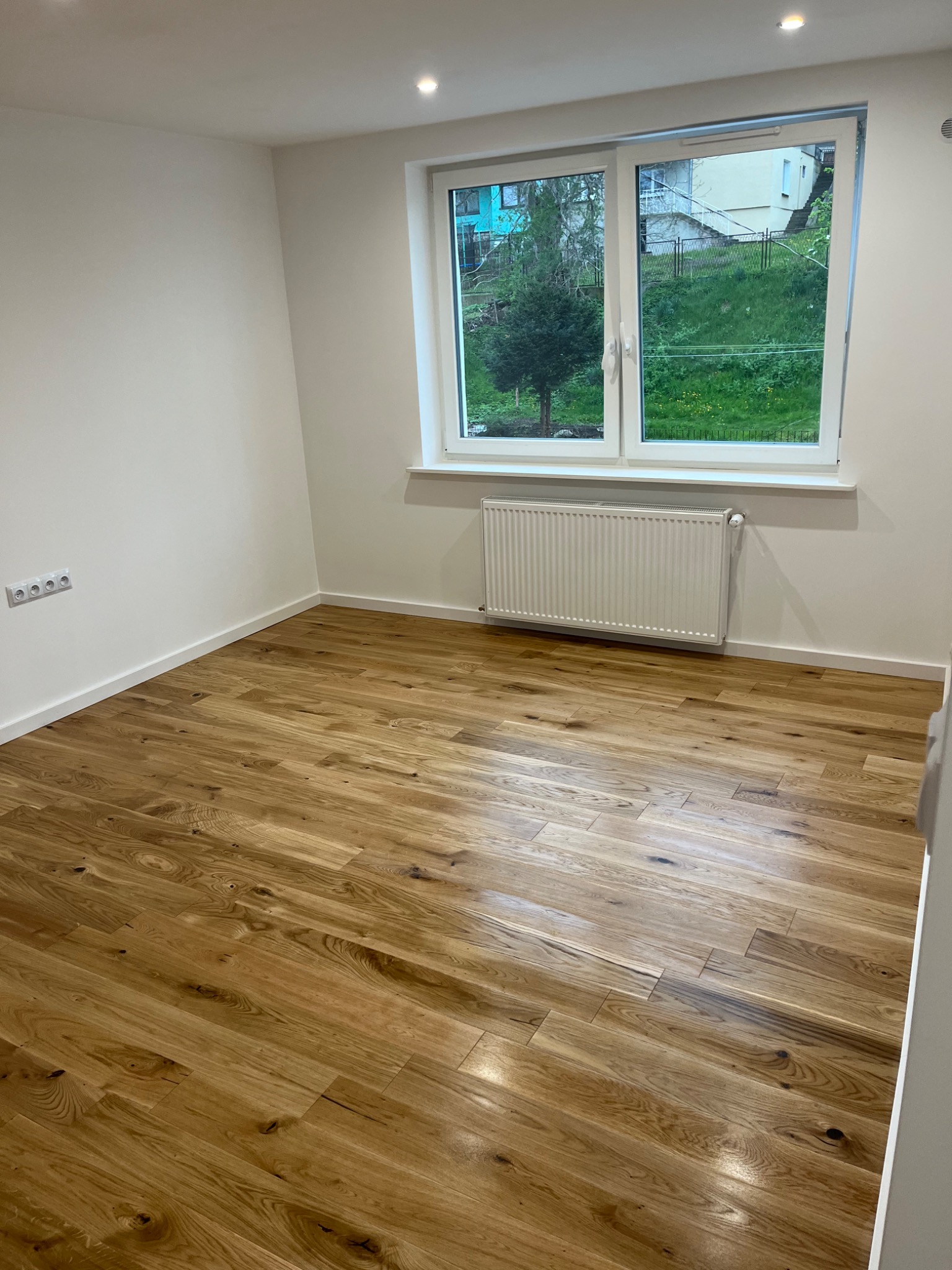 Puste, jasne pomieszczenie z drewnianą podłogą, białymi ścianami, grzejnikiem pod oknem z widokiem na zieleń i czterema gniazdkami elektrycznymi.