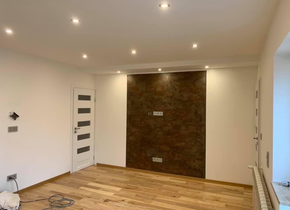 Pokój po remoncie z drewnianą podłogą, białe ściany i dekoracyjna ściana z efektem rdzy pod telewizor. Widoczne gniazdka elektryczne, oświetlenie sufitowe, białe drzwi z przeszkleniami i grzejnik...