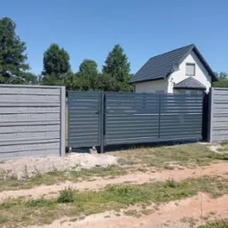 Nowoczesne ogrodzenie posesji z przesuwną bramą w kolorze grafitowym, połączone z betonowymi panelami, na tle domu jednorodzinnego i zieleni.