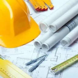 Żółty kask budowlany, poziomica, miara zwijana, kołki rozporowe i zwinięte plany architektoniczne ułożone na rysunku technicznym.