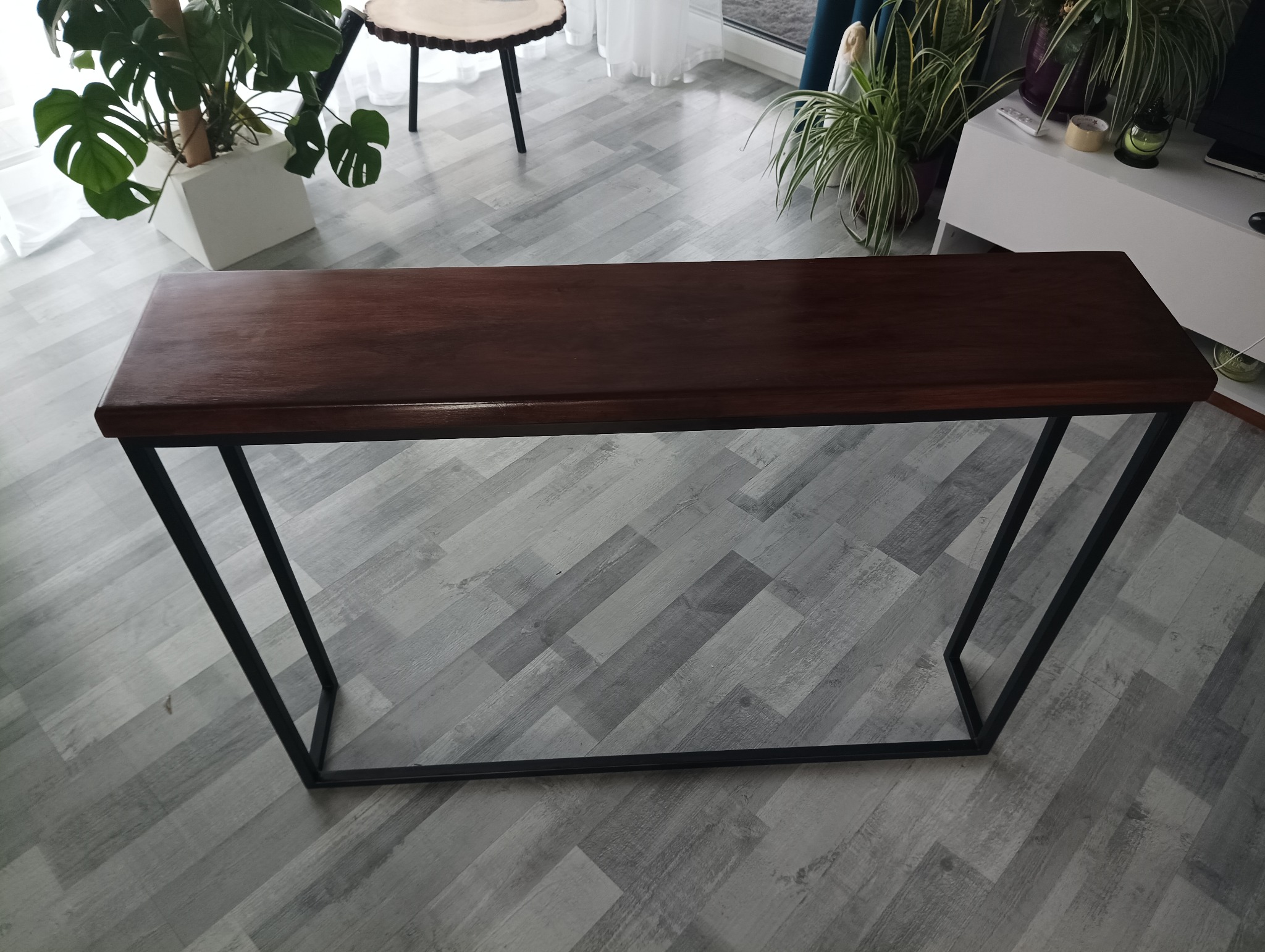 Nowoczesny stół konsola z ciemnobrązowym blatem i czarnymi metalowymi nogami, ustawiony na szarych panelach podłogowych w jasnym wnętrzu z roślinami doniczkowymi w tle.