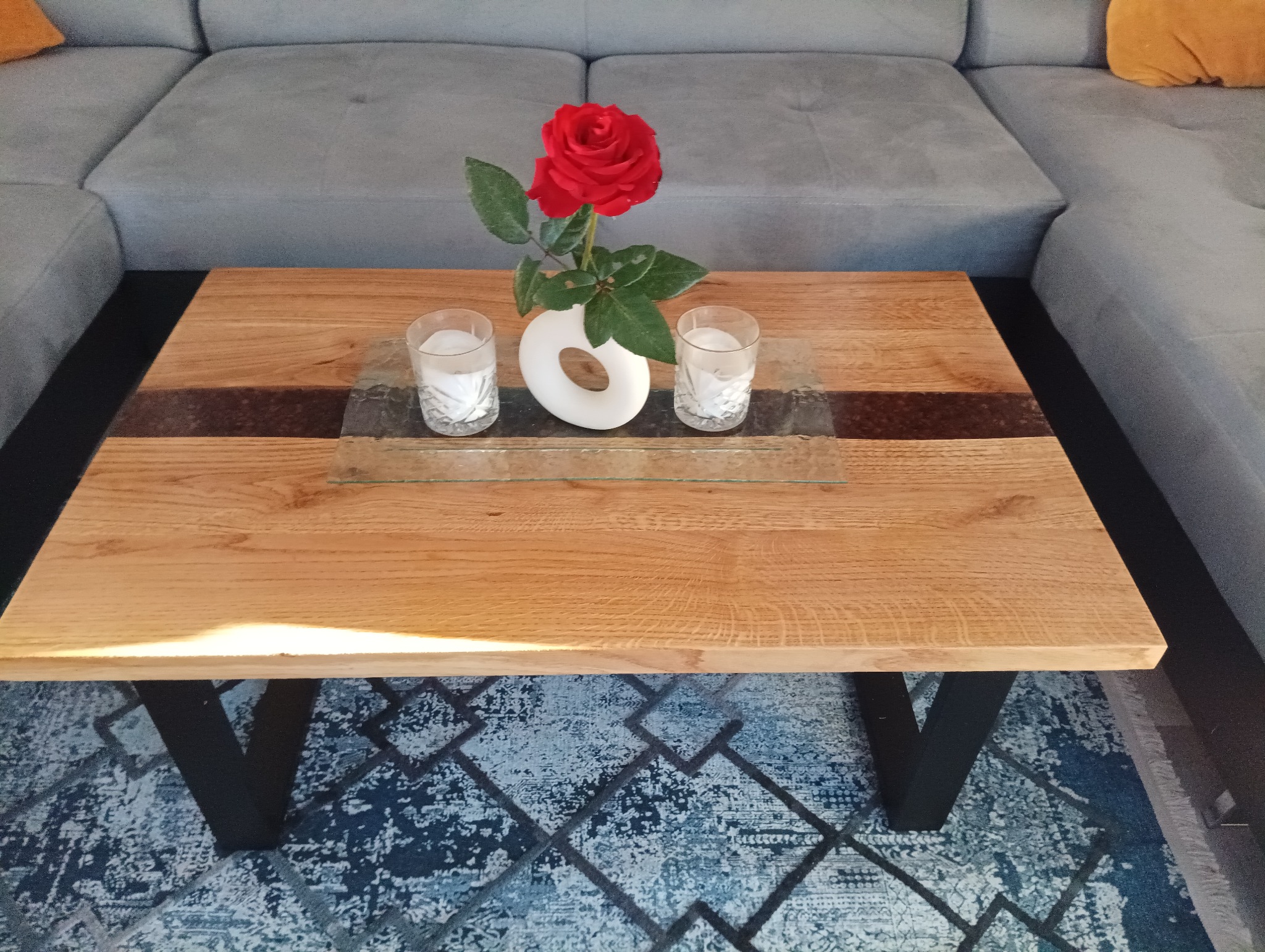 Drewniany stolik kawowy z żywicą epoksydową, ozdobiony czerwoną różą w białym wazonie i dwoma świecami w szklanych świecznikach, na tle szarej kanapy i wzorzystego dywanu.