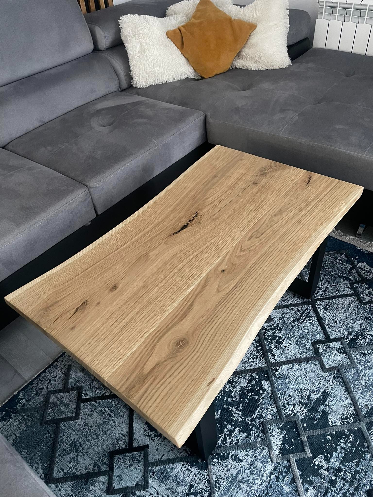 Stolik kawowy z blatem z jasnego drewna o nieregularnych krawędziach, na czarnych metalowych nogach, ustawiony na wzorzystym dywanie, obok szarej sofy z poduszkami.