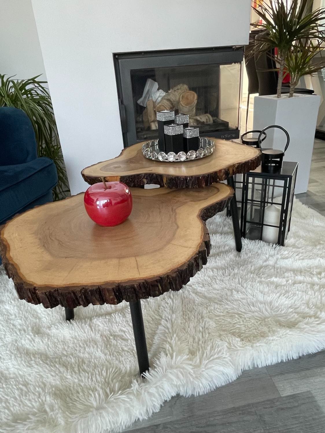 Dwu-poziomowy stolik kawowy z blatami z surowego drewna na czarnych metalowych nogach, na puszystym białym dywanie, z dekoracjami: czerwone jabłko, świeczniki i lampion. W tle kominek i rośliny...