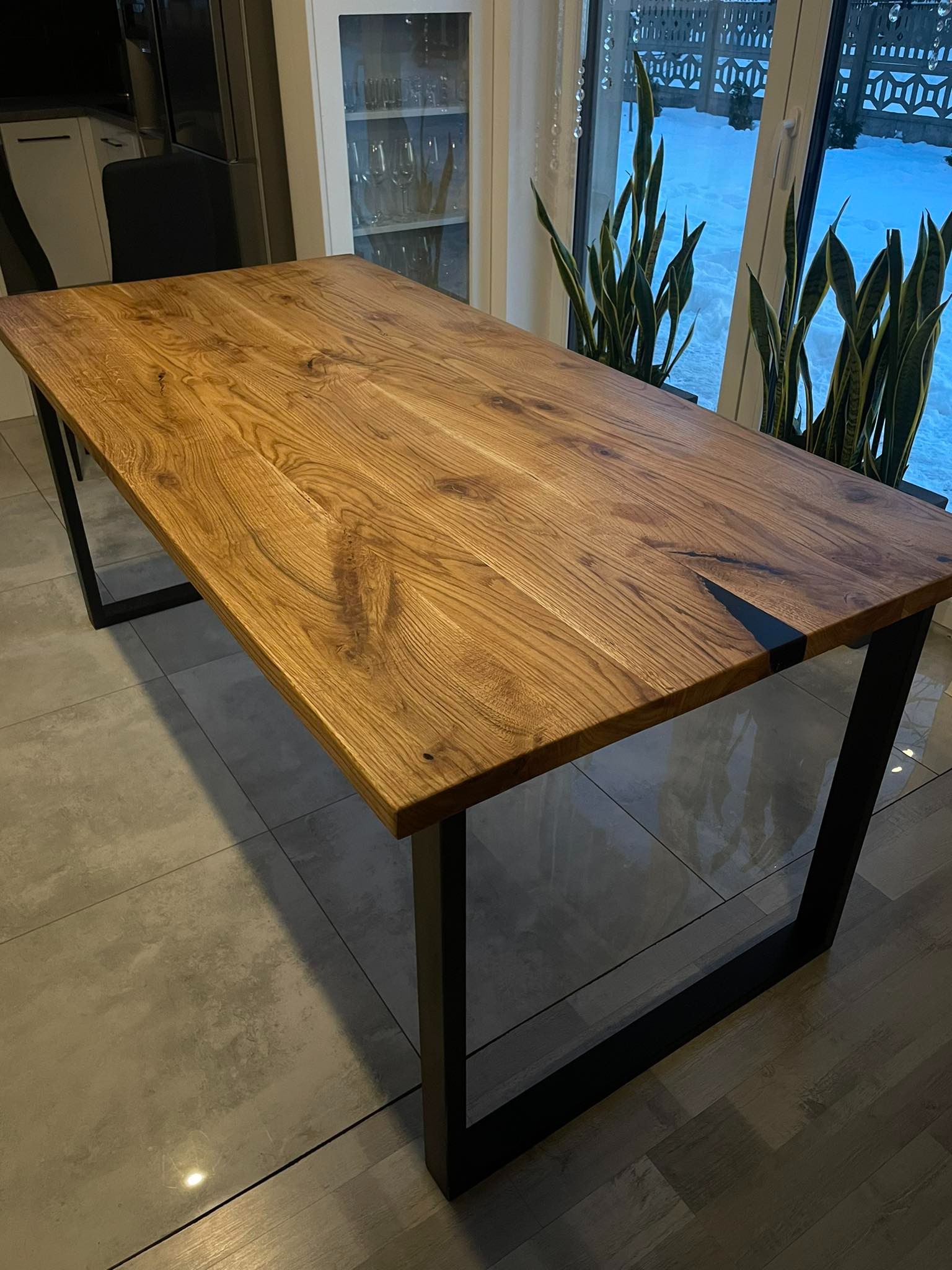 Stół z blatem z litego drewna dębowego z czarnymi, metalowymi nogami w nowoczesnym wnętrzu, widok na okno z roślinami i zaśnieżony krajobraz.