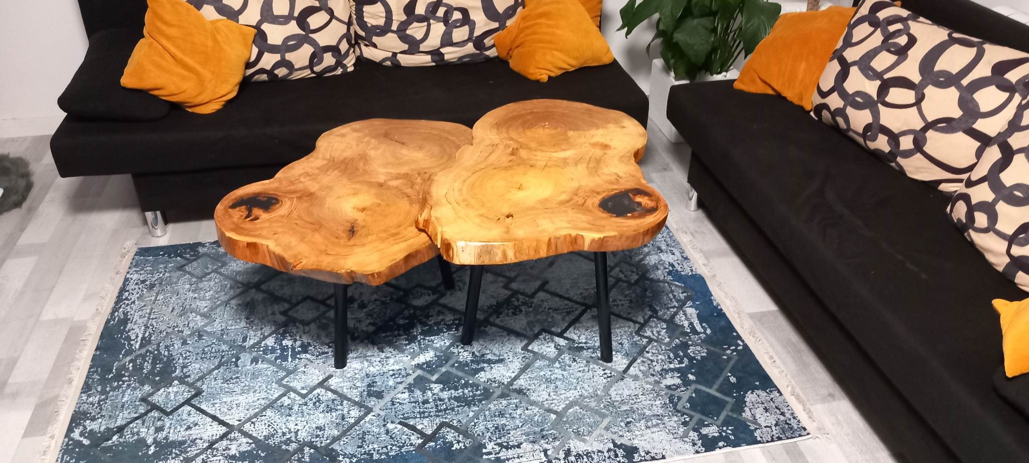 Stolik kawowy z blatem z dwóch plastrów drewna o nieregularnym kształcie, z czarnymi żywicznymi wypełnieniami, na metalowych nóżkach, na tle czarnej sofy z pomarańczowymi poduszkami i wzorzystym...
