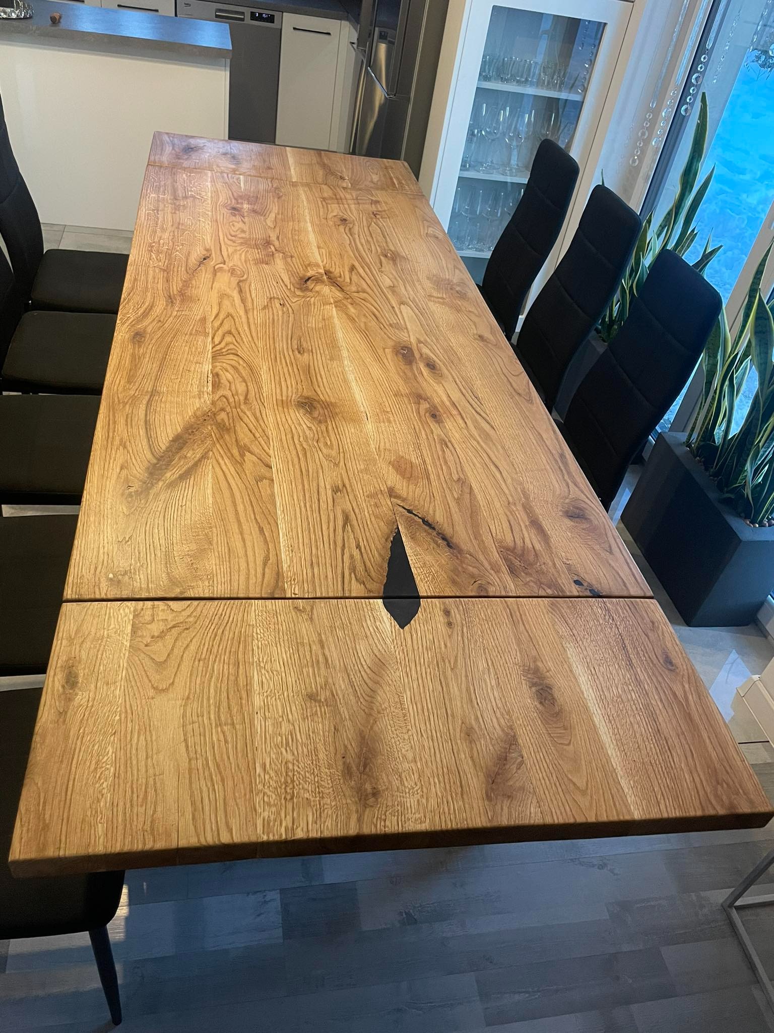 Rozkładany stół z blatem z jasnego drewna dębowego z czarną żywicą epoksydową w szczelinie, otoczony czarnymi krzesłami w nowoczesnej jadalni.