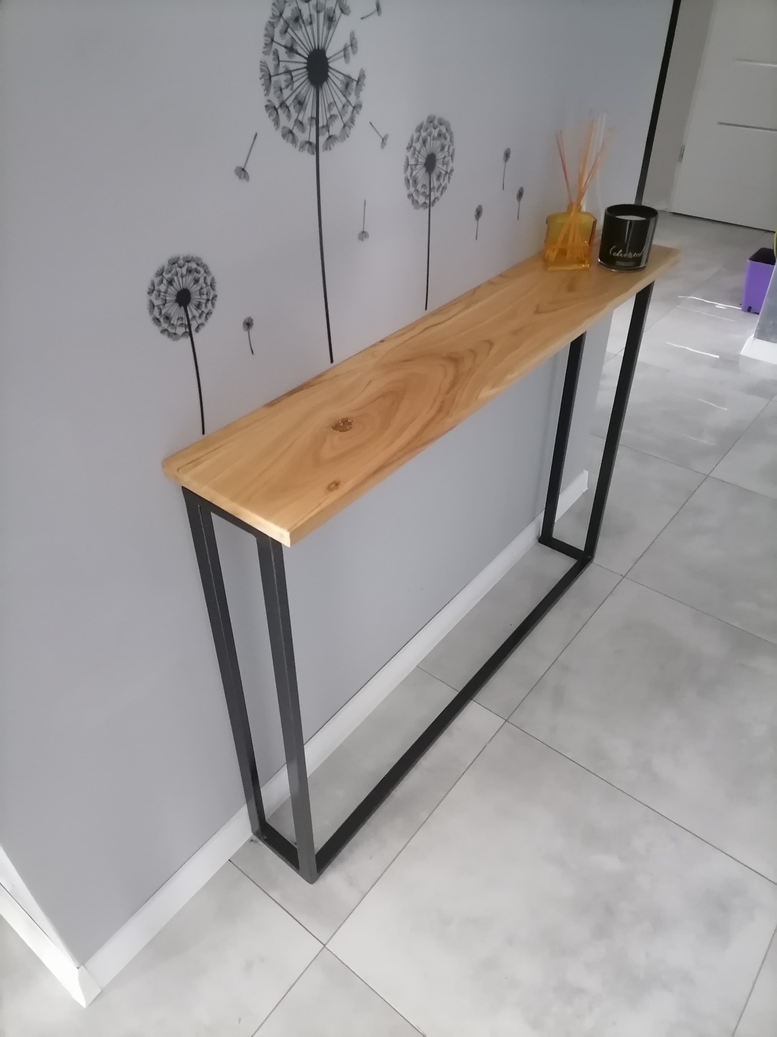 Wąski, drewniany blat konsoli z czarnymi, metalowymi nogami stoi przy szarej ścianie z naklejkami dmuchawców w Przyrowie. Na blacie dekoracyjny dyfuzor zapachowy i świeca.