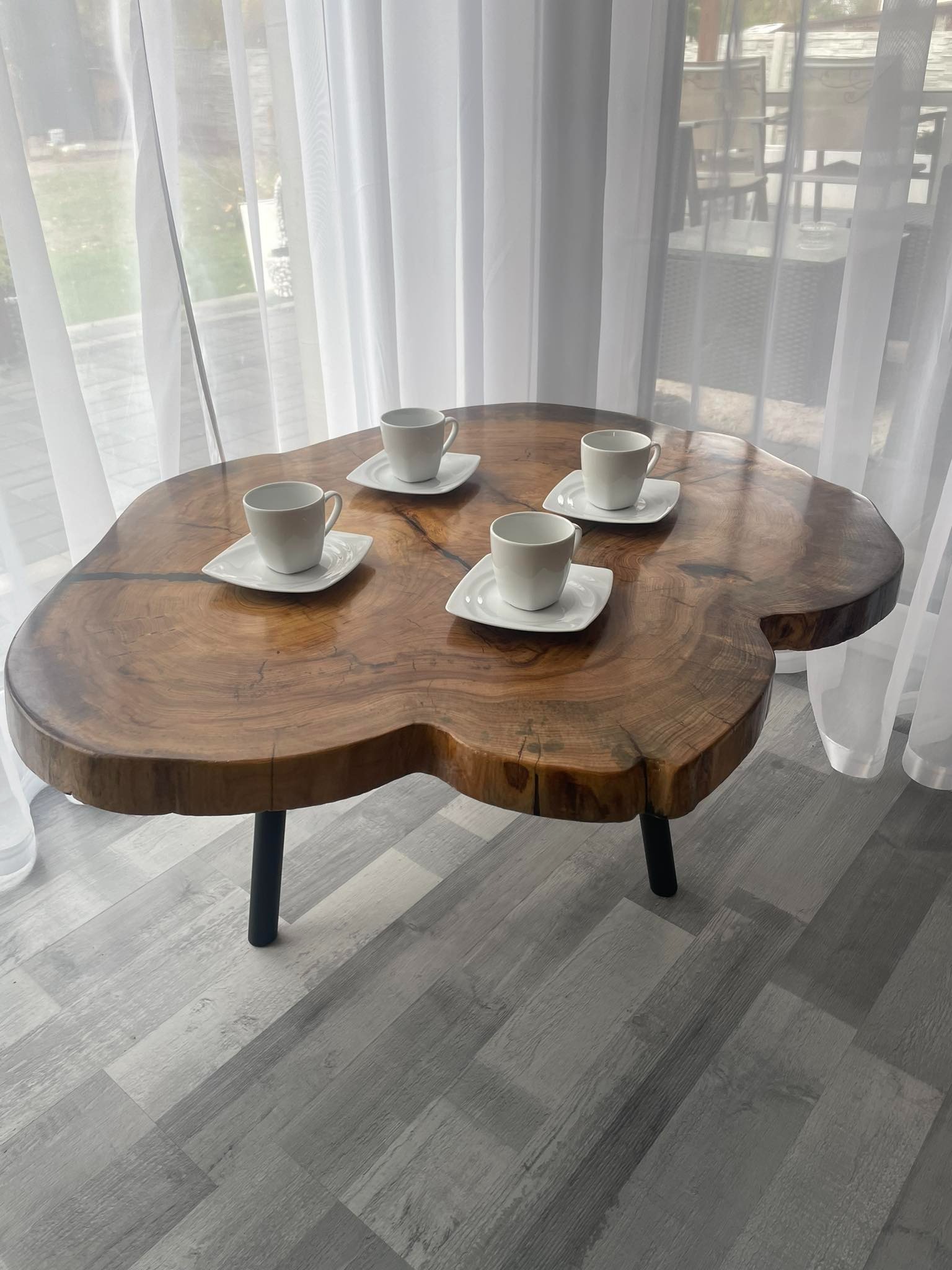 Stolik kawowy z blatem z litego drewna o nieregularnym kształcie, z widocznymi słojami i żywicą, ustawiony na czterech czarnych nogach, na blacie cztery białe filiżanki na białych podstawkach, tło...