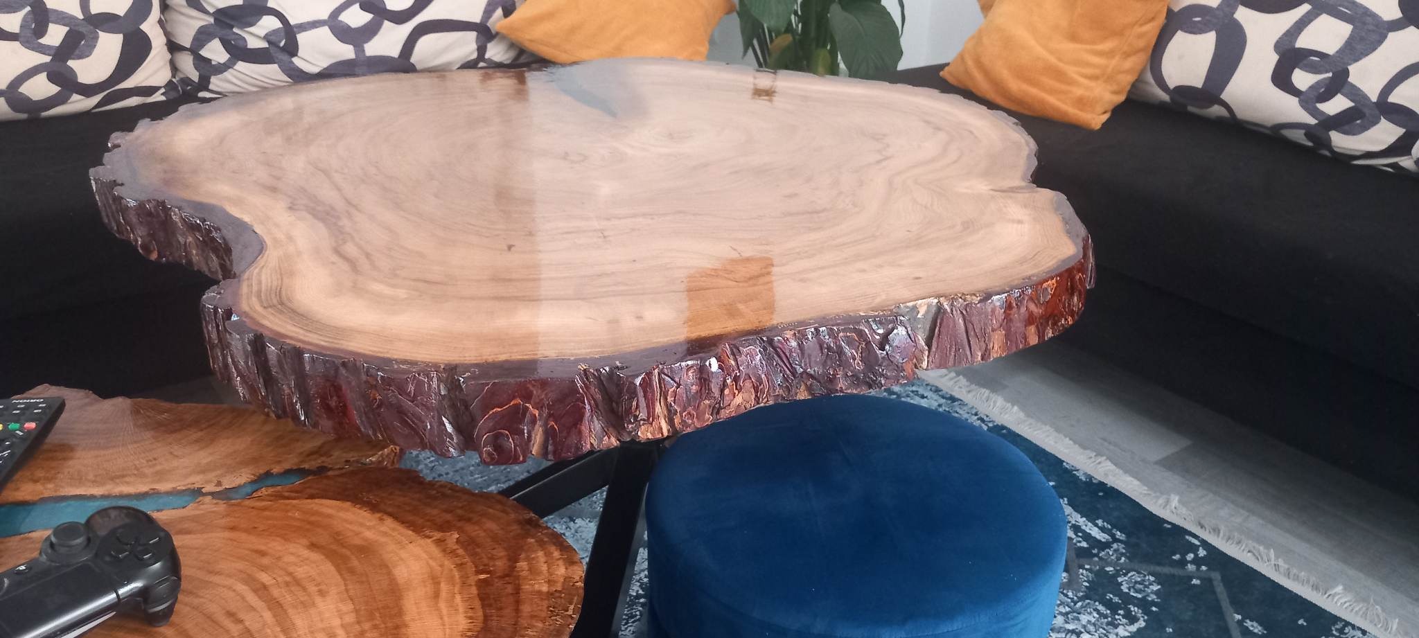 Stolik kawowy z blatem z naturalnego drewna, pokrytym żywicą, z zachowaną korą, na tle sofy z poduszkami i niebieskiego pufa.