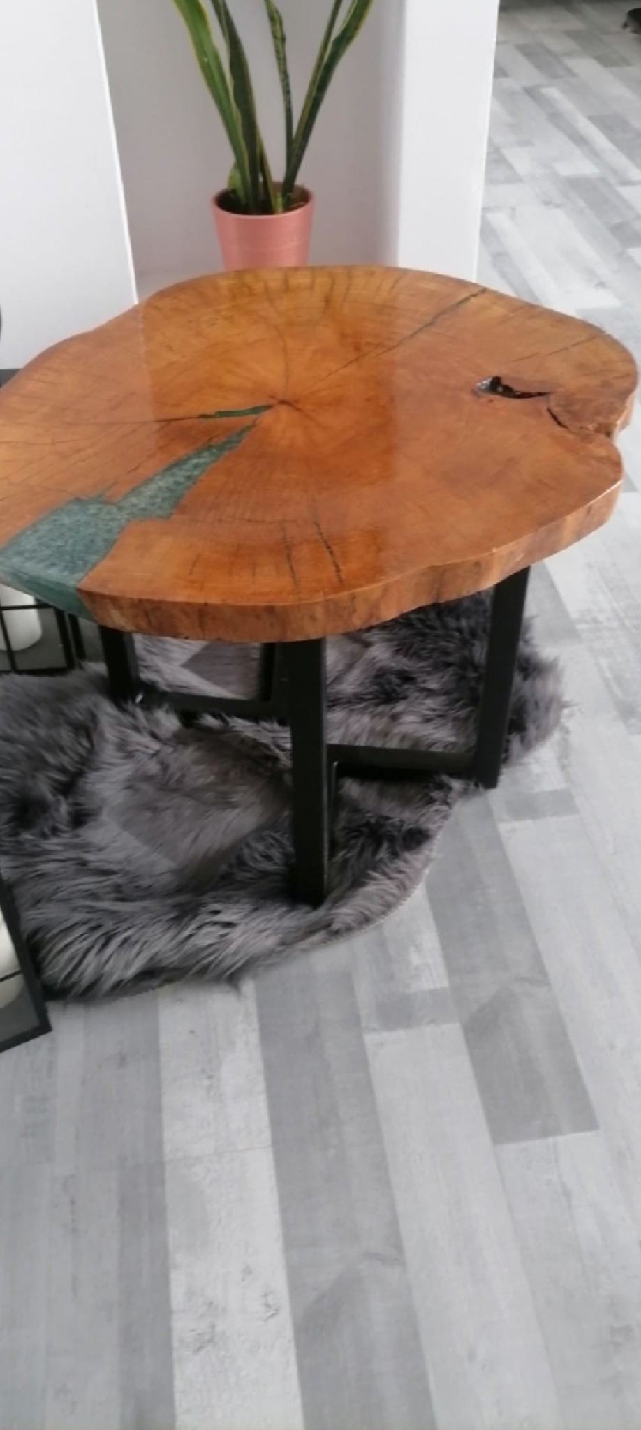 Stolik kawowy z blatem z plastra drewna i żywicy epoksydowej, czarne metalowe nogi, szary dywanik z długim włosiem, w tle roślina doniczkowa.
