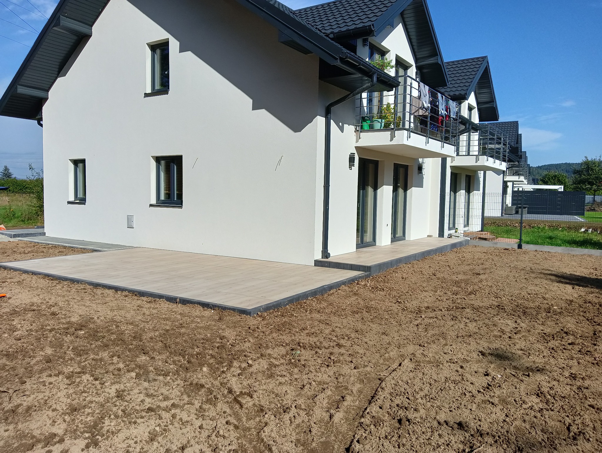 Nowoczesny taras z jasnych płyt, obrzeża z ciemnej kostki, na tle białego domu z balkonem i przygotowanego pod trawnik terenu.