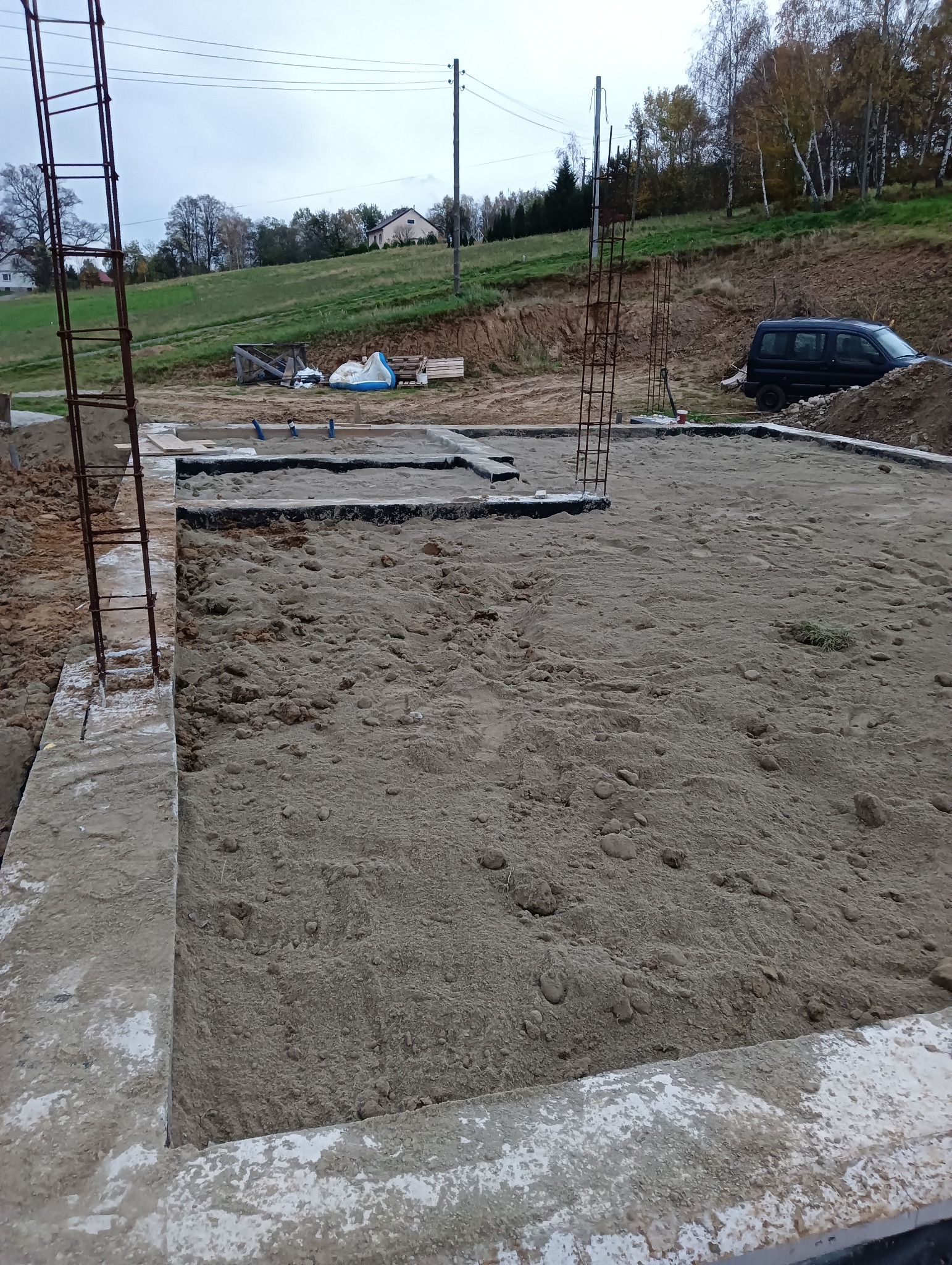 Przygotowanie fundamentów pod budowę: zalany betonowy obrys z wystającymi prętami zbrojeniowymi, teren wyrównany piaskiem, w tle dom na wzgórzu i zaparkowany samochód.
