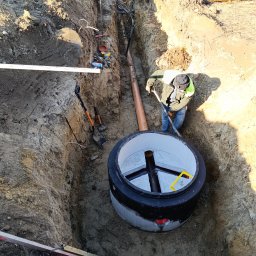 First-Bud Grzegorz Onak - Głęboki wykop ziemny z ułożoną rurą kanalizacyjną i betonowym kręgiem studni; robotnik z łopatą poprawia ułożenie, widoczne narzędzia i poziomica.