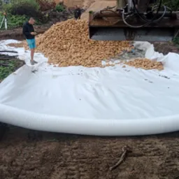 Koparka wysypuje żwir na białą geowłókninę, przygotowanie podłoża pod budowę, widoczni pracownicy i niwelator laserowy.