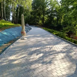 Długa, szara ścieżka z kostki brukowej wijąca się przez park, obok drzew i zieleni. Widoczny parasol i pojazd w oddali, oświetlone słonecznym światłem.