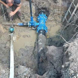 First-Bud Grzegorz Onak - Instalacja wodociągowa w wykopie, z widocznym pracownikiem i fragmentem drabiny. Rury w kolorze niebieskim, teren wokół błotnisty. Prace ziemne w toku.