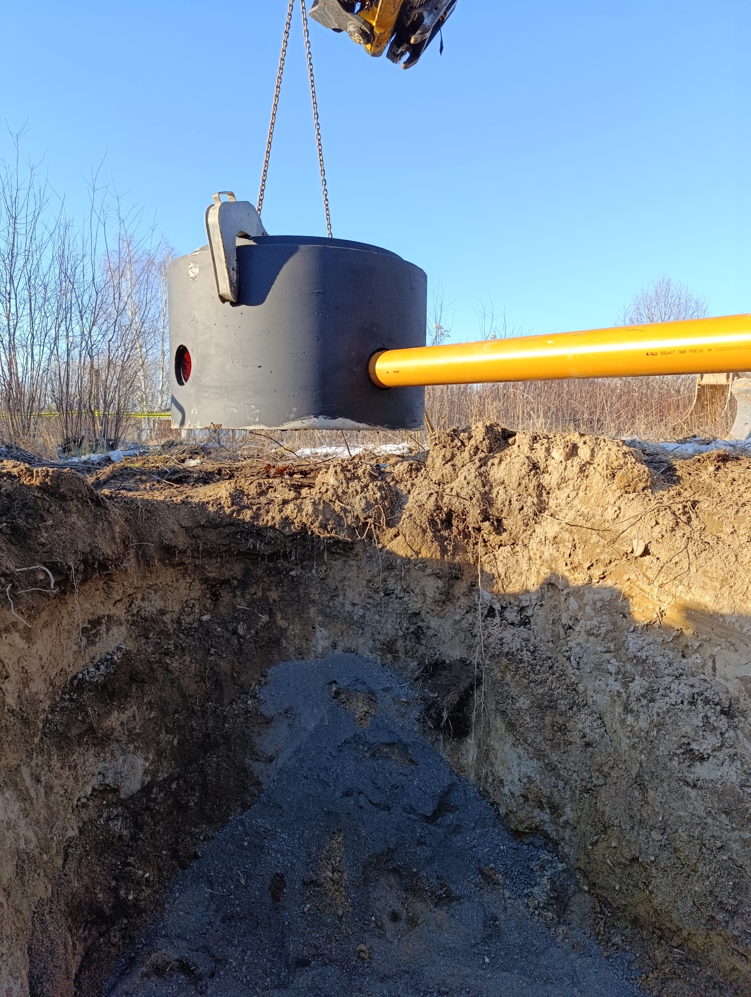 Montaż betonowego zbiornika z żółtą rurą odpływową w wykopie ziemnym, podwieszony na łańcuchach przez maszynę budowlaną, widok na ziemię i błękitne niebo.