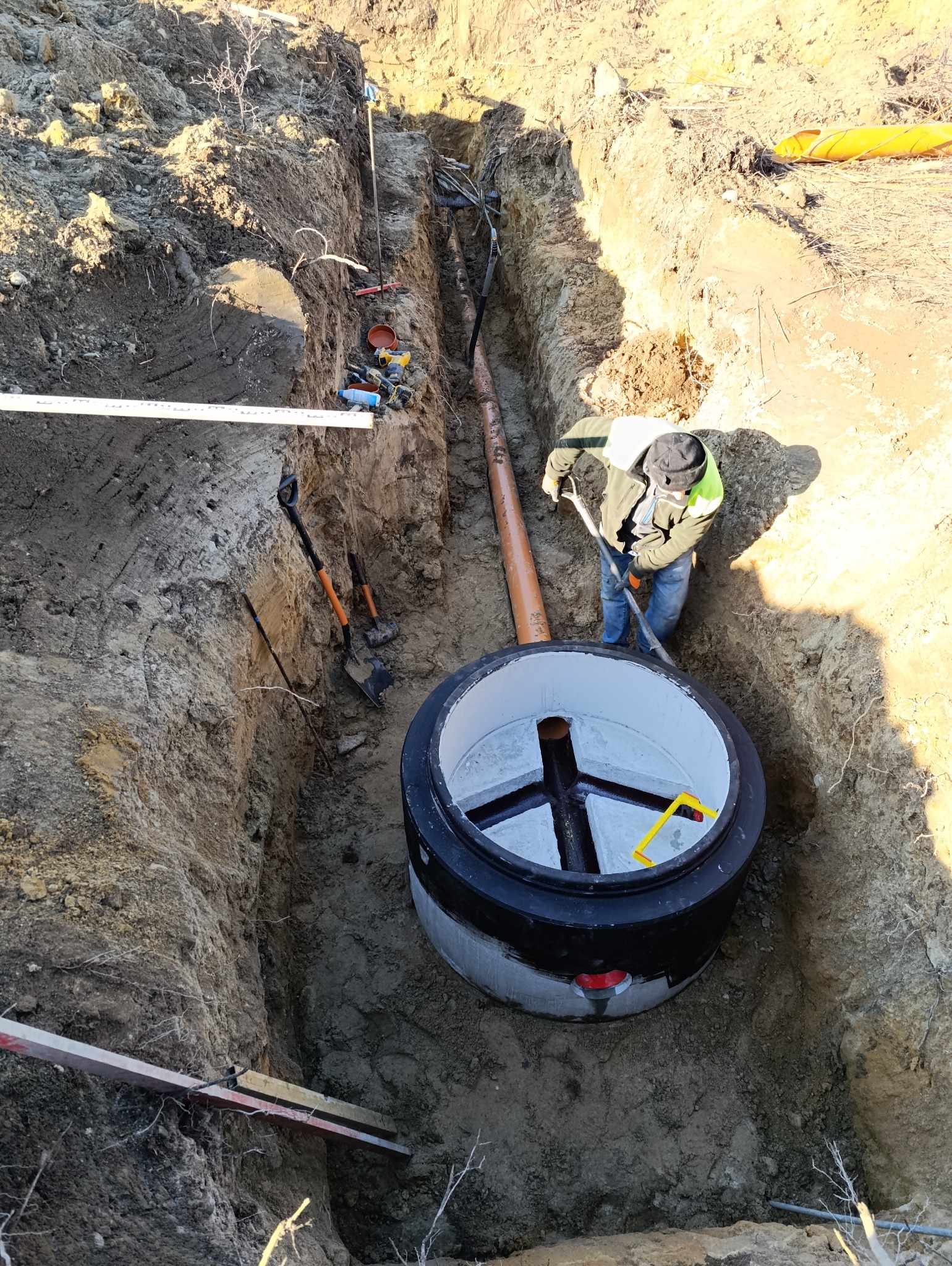 Głęboki wykop ziemny z ułożoną rurą kanalizacyjną i betonowym kręgiem studni; robotnik z łopatą poprawia ułożenie, widoczne narzędzia i poziomica.