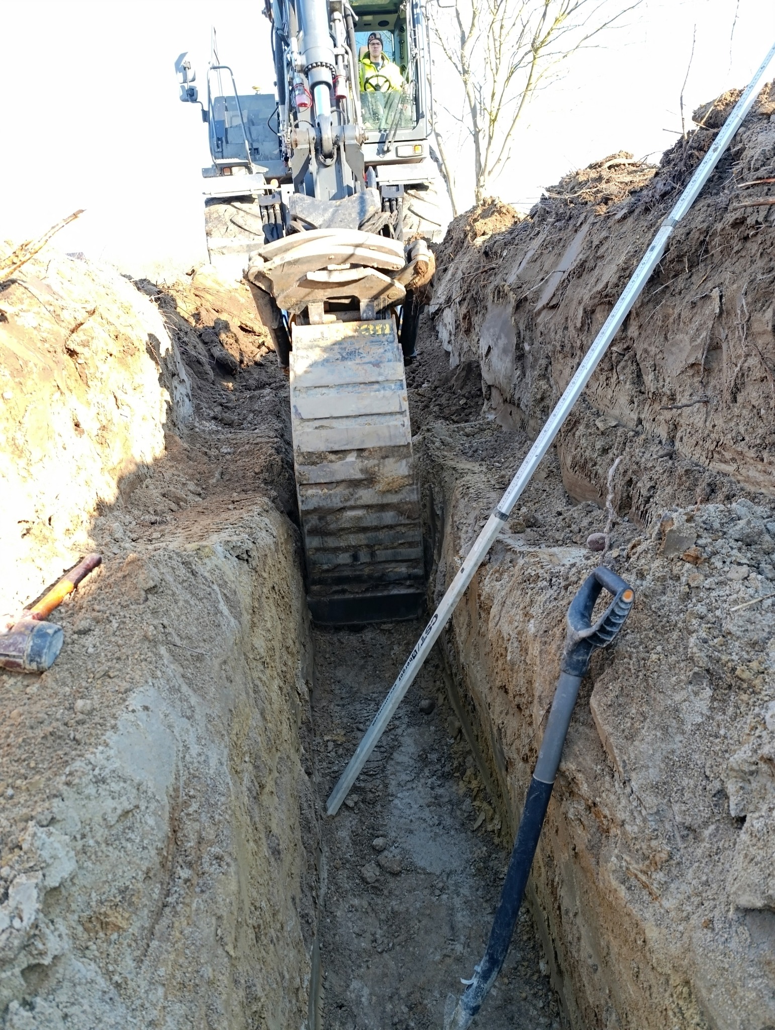 Wąski wykop w ziemi z widoczną łyżką koparki i operatorem w kabinie, obok leży długa poziomica i wbita jest łopata.