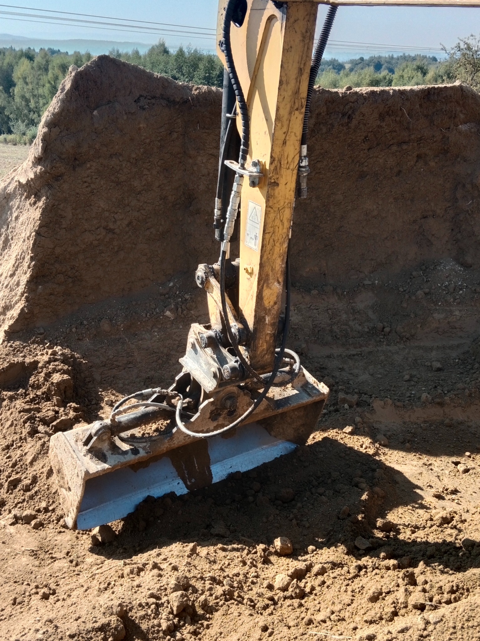 Żółta koparka z łyżką zagłębiająca się w brązową ziemię, w tle kopiec ziemi i zielone drzewa.