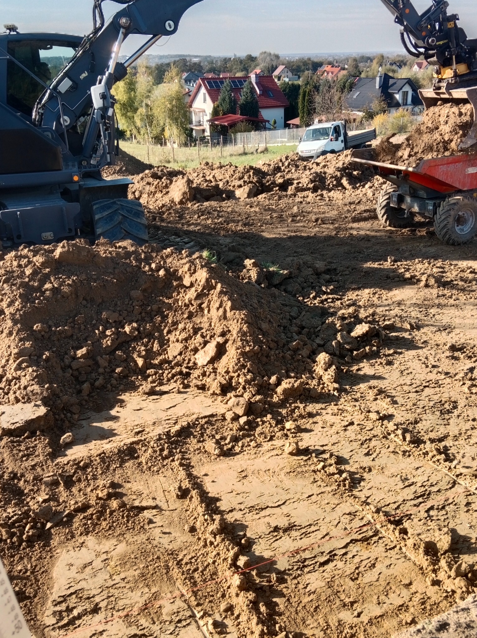 Dwie koparki w trakcie prac ziemnych na placu budowy, w tle zabudowa jednorodzinna i biały samochód dostawczy.