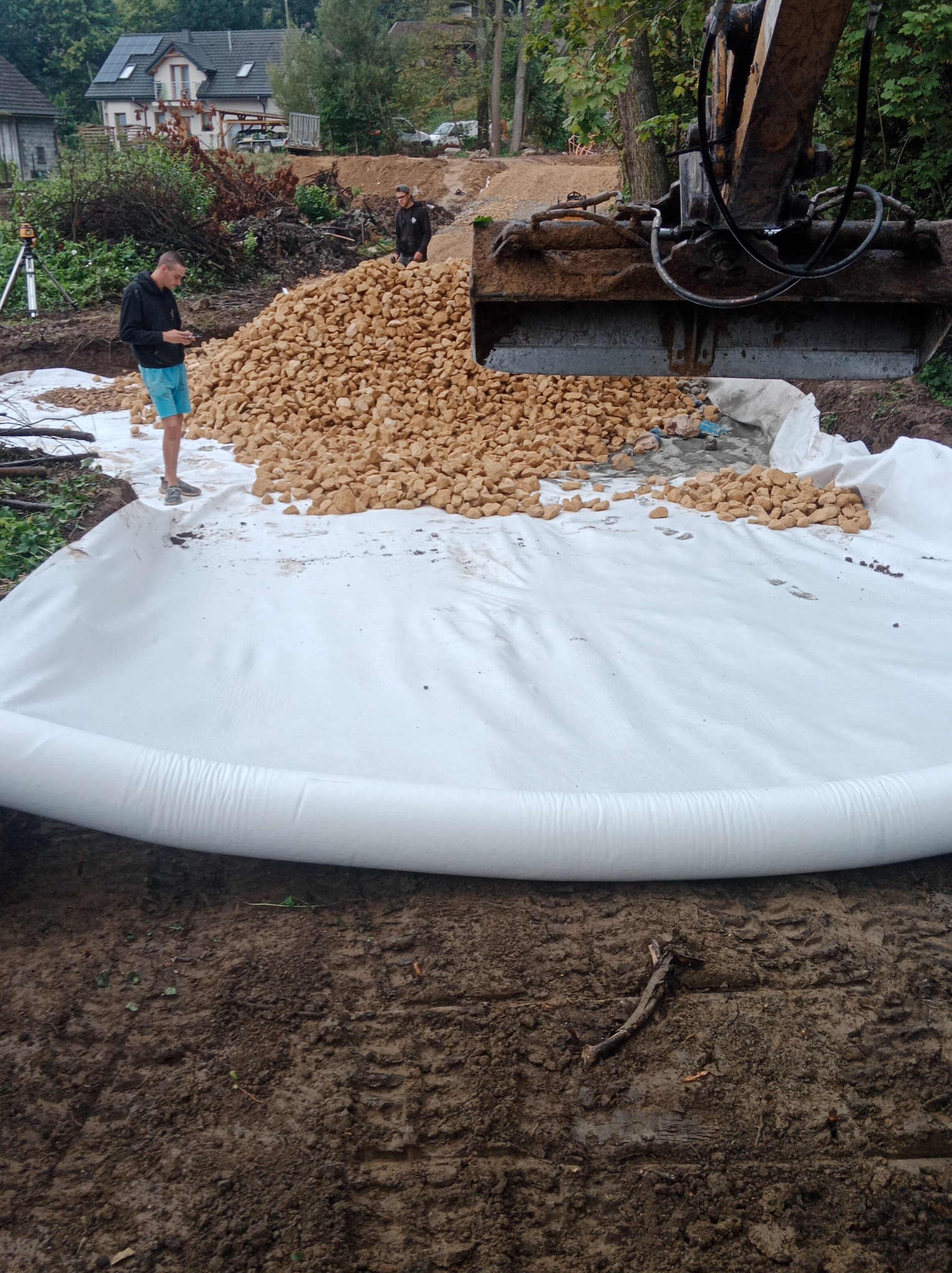 Koparka wysypuje żwir na białą geowłókninę, przygotowanie podłoża pod budowę, widoczni pracownicy i niwelator laserowy.
