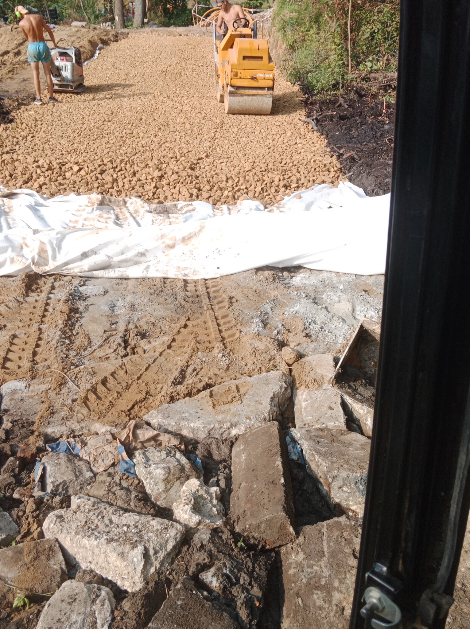 Drogowa walcarka ubija nawierzchnię z kruszywa na przygotowanym podłożu, widoczny fragment białej geowłókniny i pozostałości po rozbiórce, dwóch pracowników w tle.