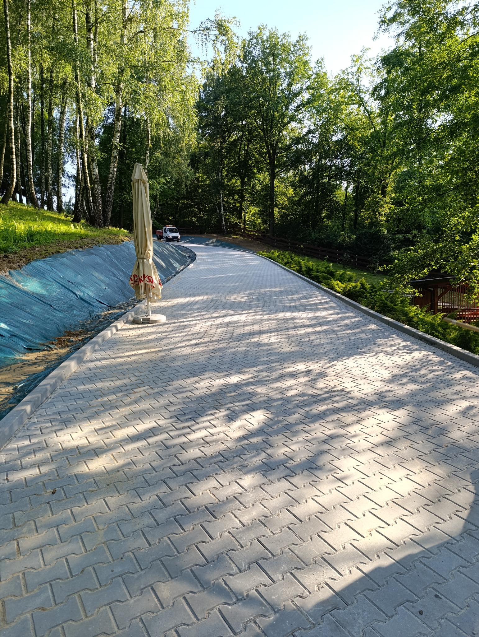 Długa, szara ścieżka z kostki brukowej wijąca się przez park, obok drzew i zieleni. Widoczny parasol i pojazd w oddali, oświetlone słonecznym światłem.
