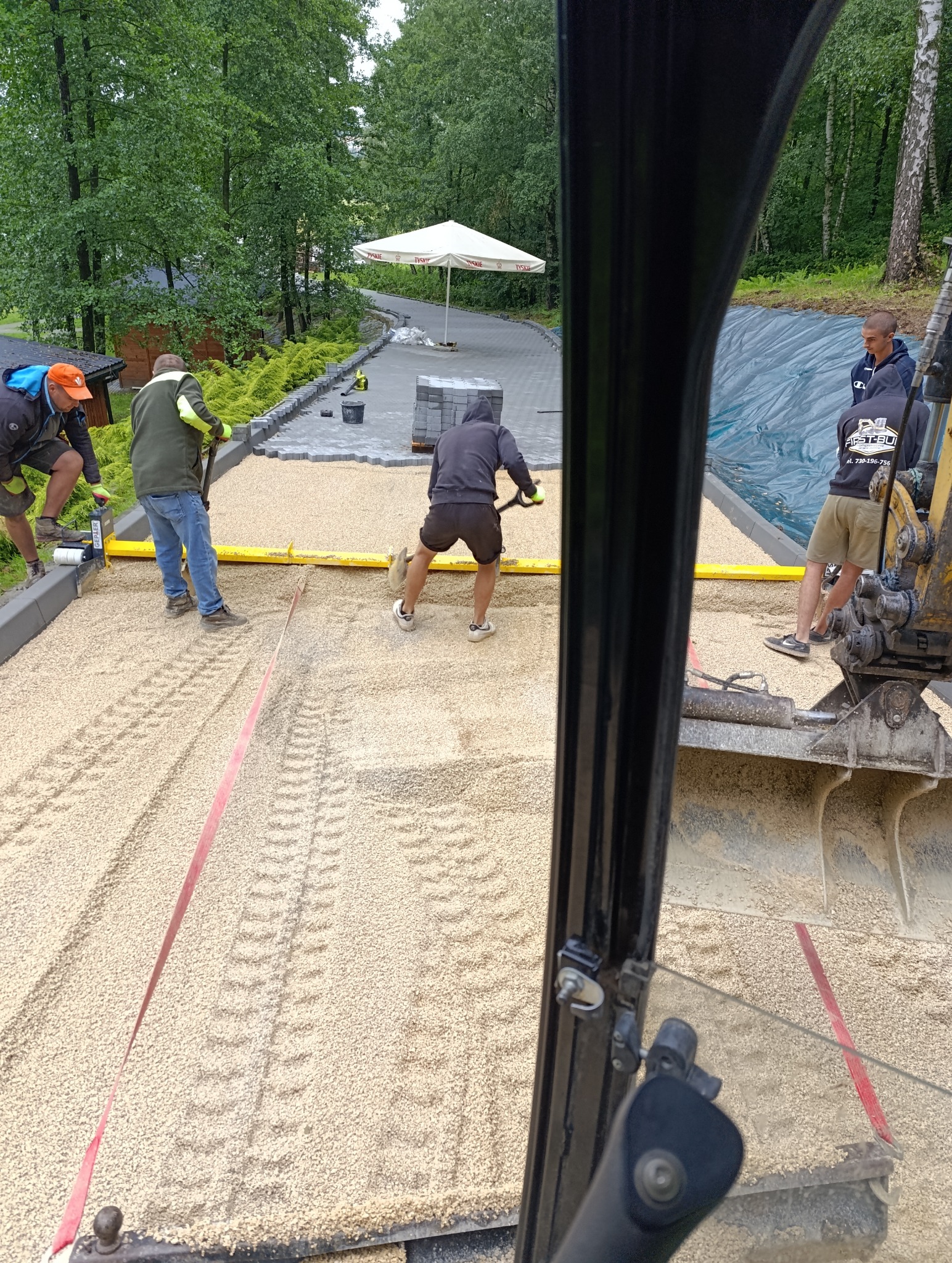 Ekipa robotników wyrównuje podłoże pod kostkę brukową, widok z kabiny koparki. W tle ułożona kostka i parasol.
