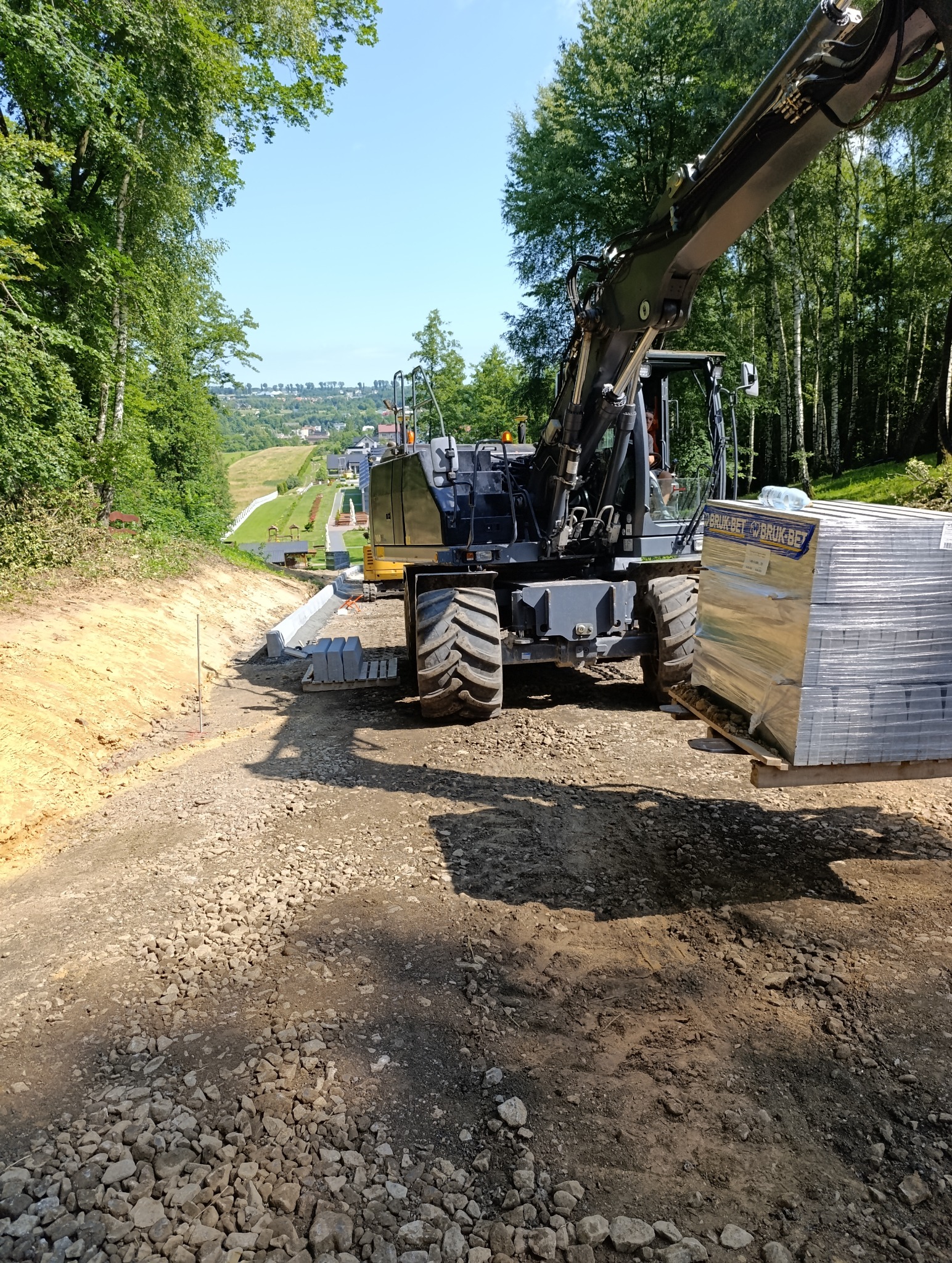Żwirowa droga w trakcie utwardzania z paletą kostki i koparką na tle zieleni i zabudowań. Widok z perspektywy ziemi, w słoneczny dzień.