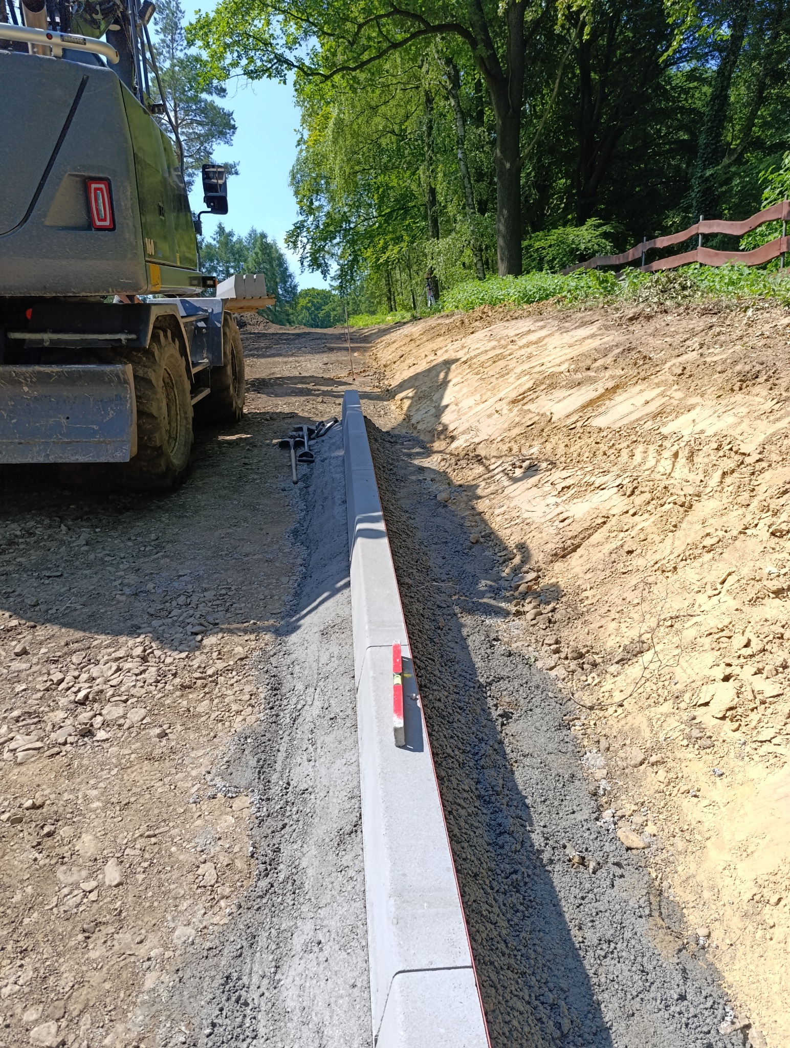 Układanie obrzeży betonowych na przygotowanym podłożu, widoczna poziomica, w tle maszyna budowlana i teren zielony. Prace brukarskie w toku.