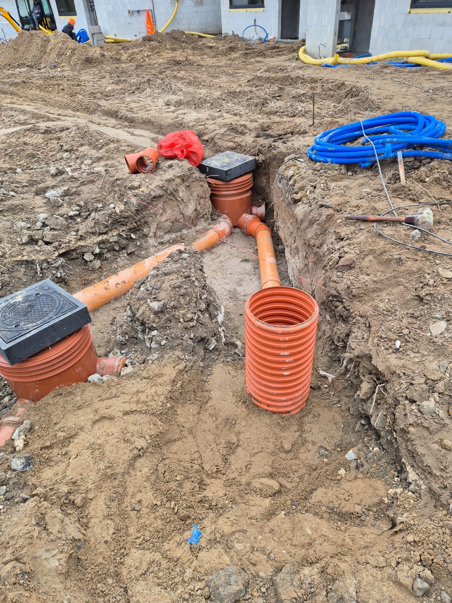 Instalacja drenażowa w wykopie: pomarańczowe rury, studzienki z czarnymi pokrywami, niebieskie rury karbowane. W tle budynek w stanie surowym i koparka.