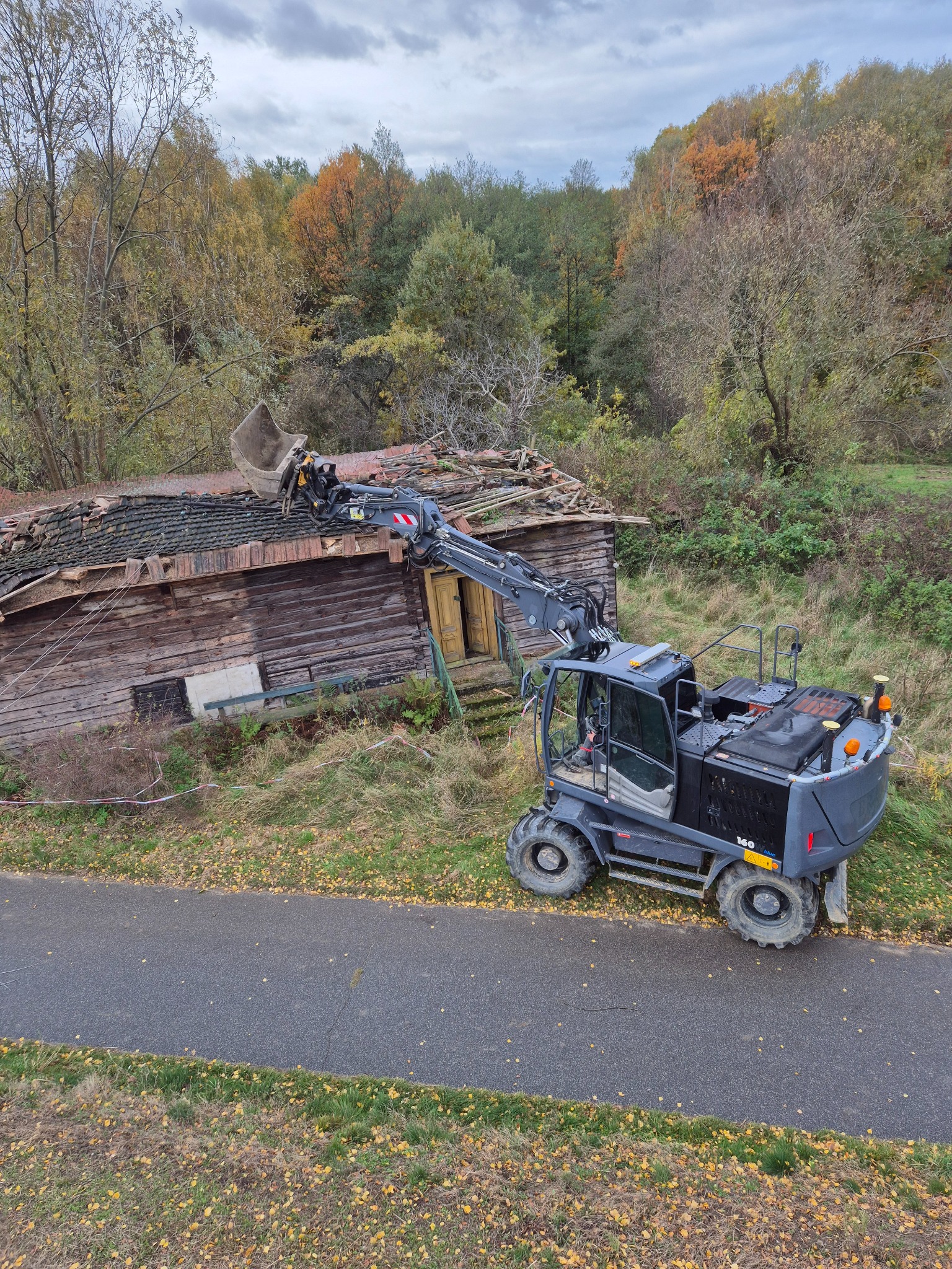 Koparka kołowa w trakcie rozbiórki drewnianego budynku z ceglanym dachem, otoczona zielenią i drzewami w jesiennej scenerii. Widok z góry na maszynę i częściowo zdemontowany budynek.
