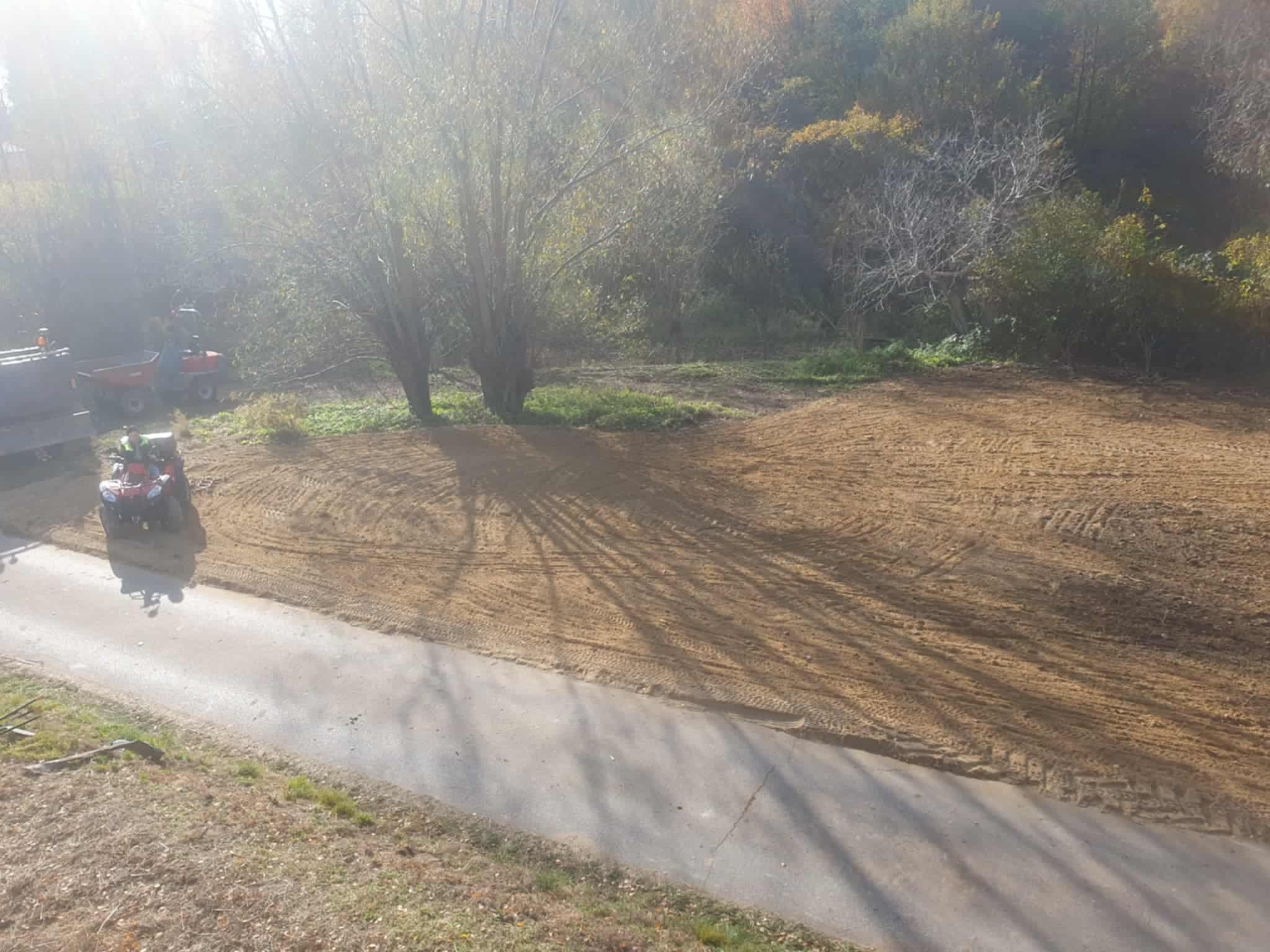 Przygotowany teren pod budowę z użyciem maszyn, widoczne ślady opon na piasku i częściowo asfaltowa droga. W tle drzewa i krzewy.