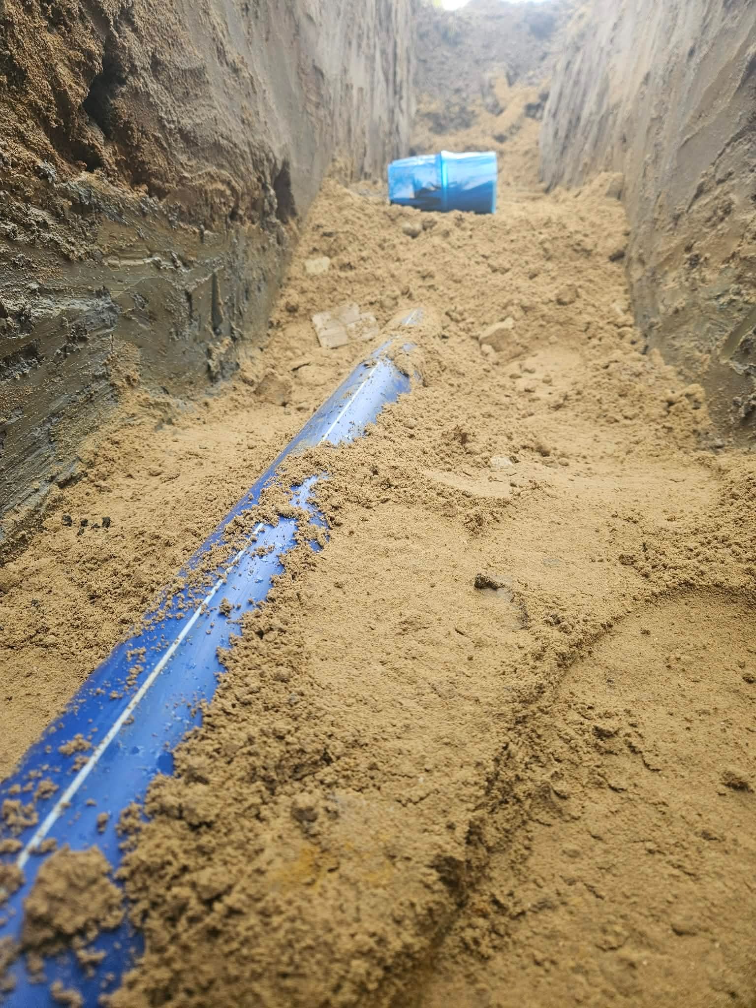 Niebieska rura w wykopie zasypana piaskiem, widoczne ściany wykopu i fragment niebieskiego elementu instalacji w tle. Ujęcie z dołu, podkreślające głębokość wykopu.