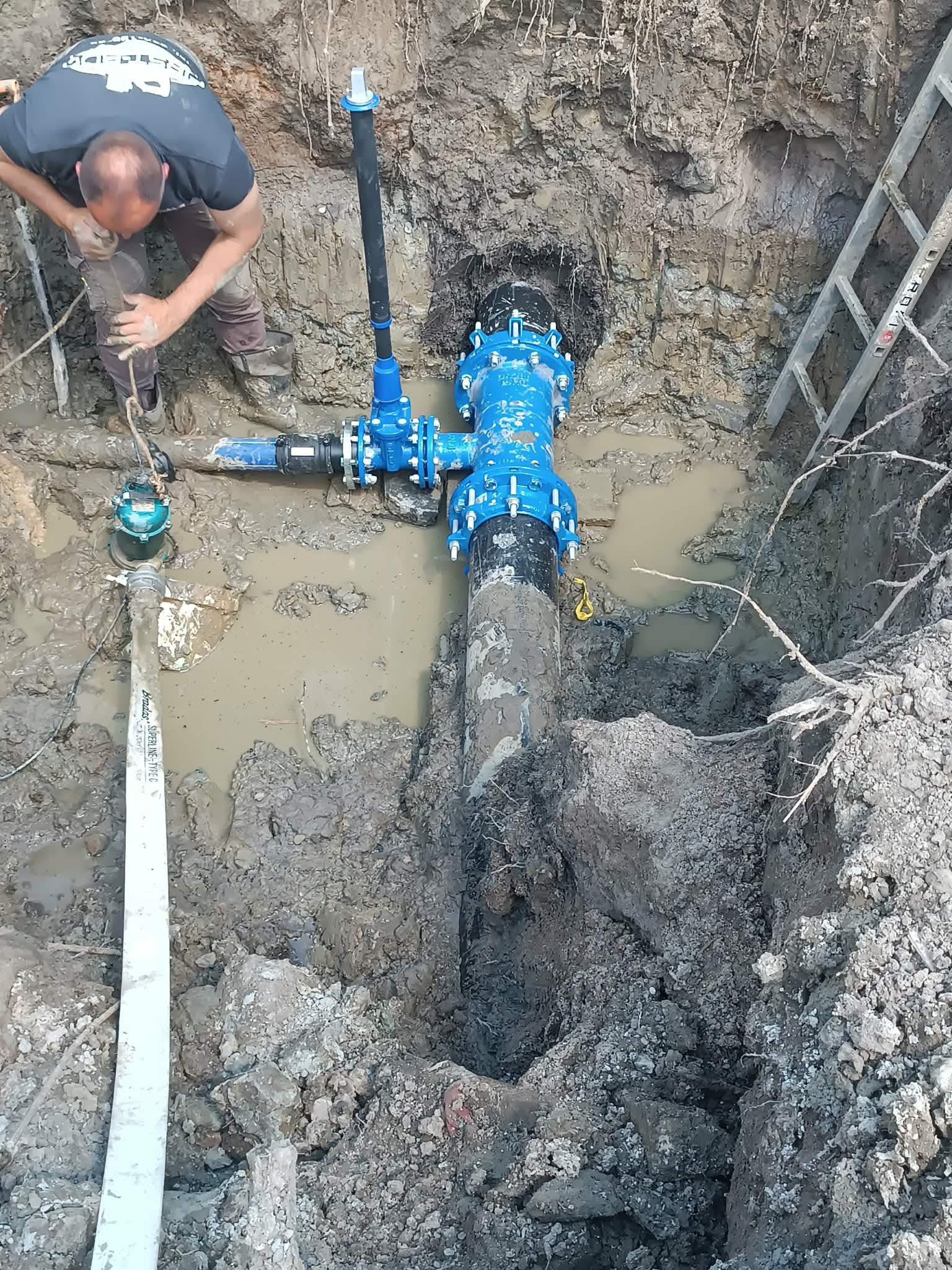 Instalacja wodociągowa w wykopie, z widocznym pracownikiem i fragmentem drabiny. Rury w kolorze niebieskim, teren wokół błotnisty. Prace ziemne w toku.