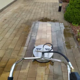 Czyszczenie kostki brukowej na podjeździe za pomocą myjki ciśnieniowej z okrągłą dyszą, widoczne mokre i czyste fragmenty kostki.