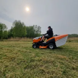 Mężczyzna w czapce i ciemnej bluzie kosi trawę traktorkiem Stihl na rozległym, częściowo skoszonym polu w pochmurny dzień.