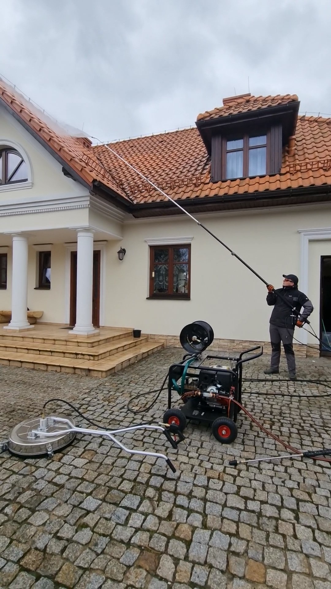 Czyszczenie dachu domu jednorodzinnego z czerwonej dachówki za pomocą myjki ciśnieniowej przez pracownika w ubraniu roboczym, na pierwszym planie maszyna czyszcząca na kółkach i dysza...